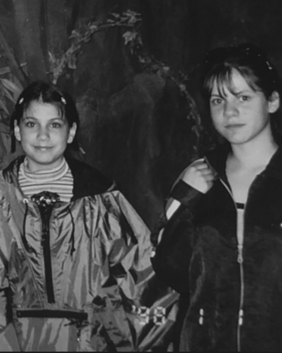 Two young girls standing next to each other in front of a large decorative plant.