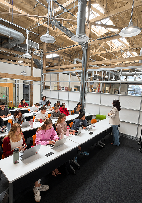 Modern office space with people working on laptops, large windows, and exposed ceiling beams.