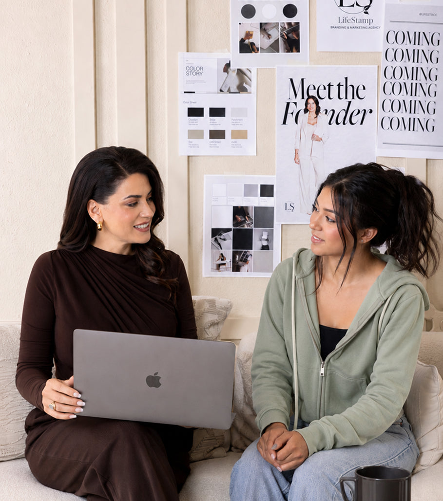 Two women sitting on a couch with a laptop, surrounded by framed posters on the wall.
