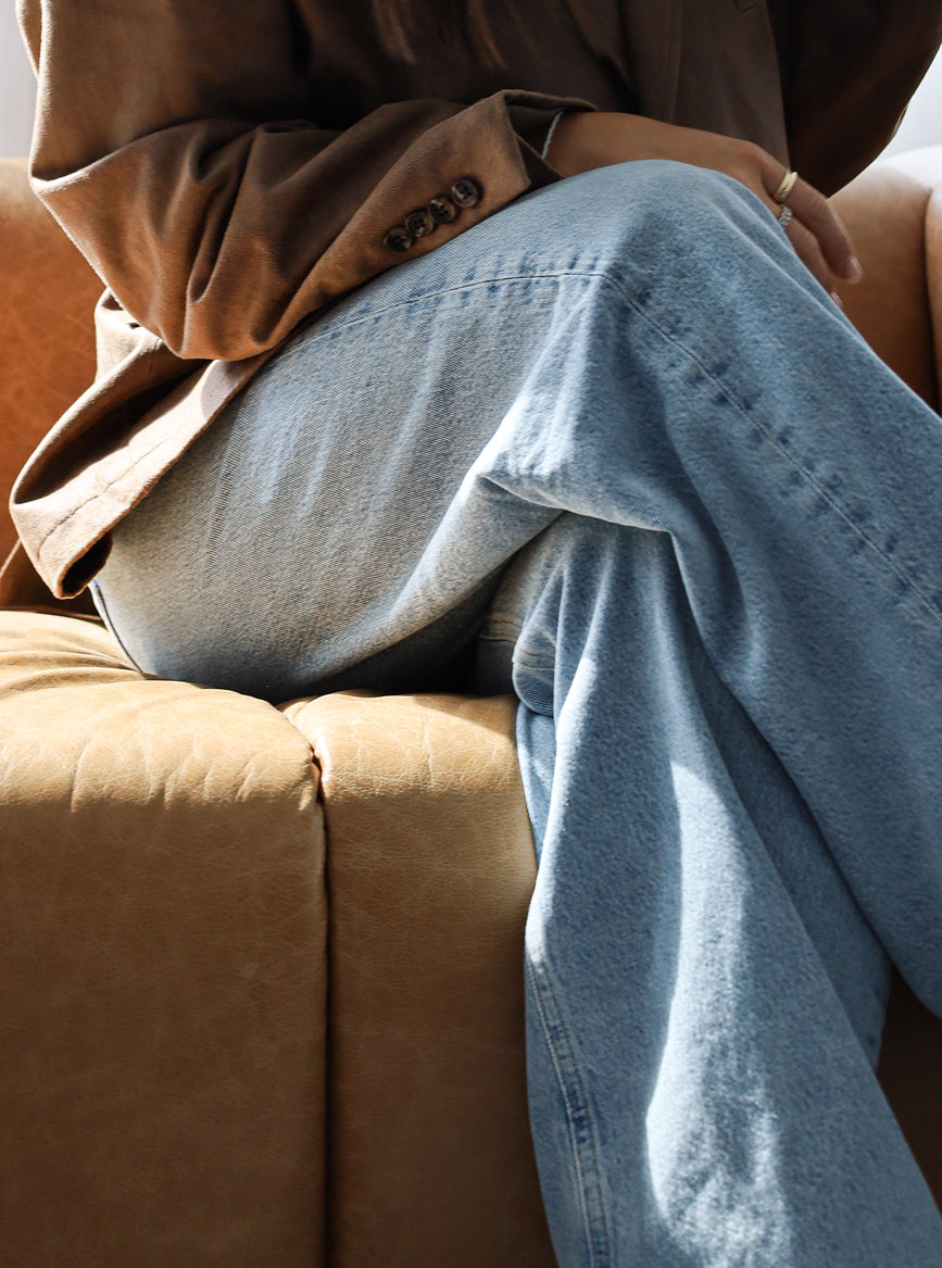 Person wearing a brown coat and blue jeans sitting on a brown couch.