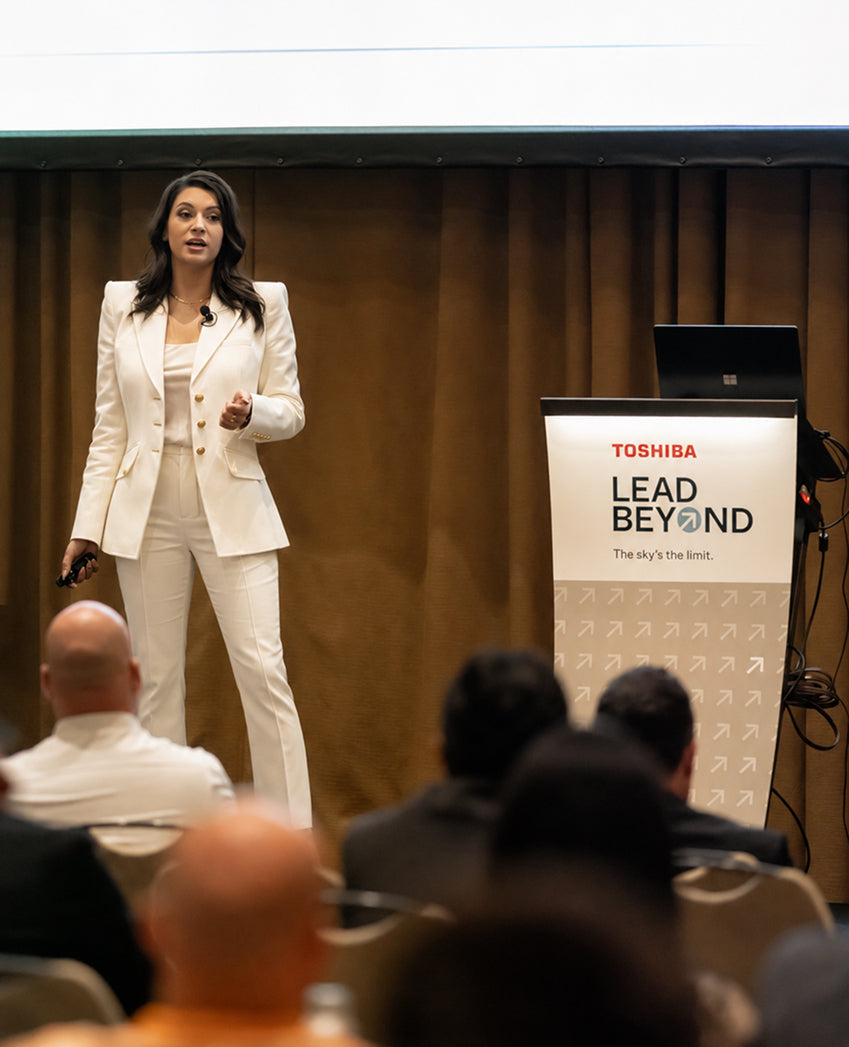 Woman in a white suit standing at a podium with Toshiba branding in an indoor setting.
