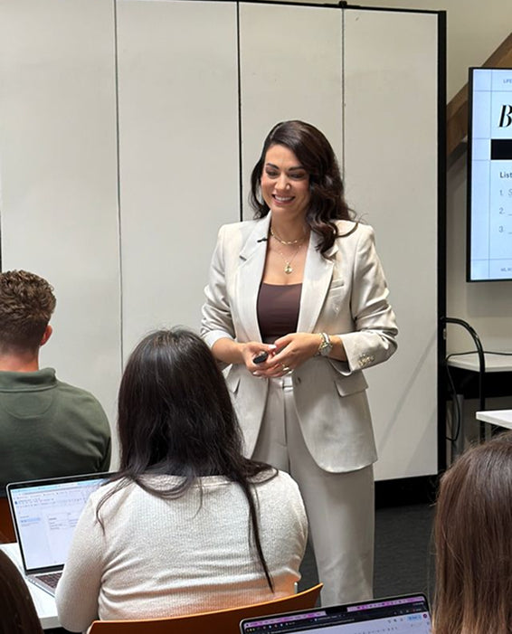 Woman in a beige suit presenting to an audience in a classroom setting.