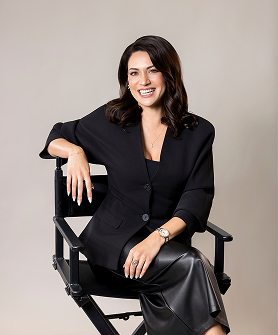 Woman in a black suit sitting on a director's chair against a plain background
