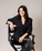Woman in a black suit sitting on a director's chair against a plain background