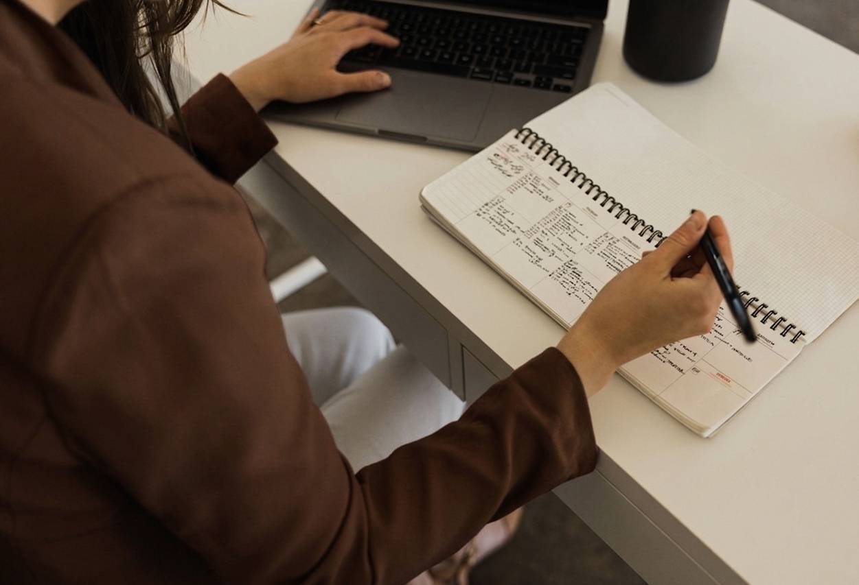 Person working at a desk with a laptop and notebook, taking notes.